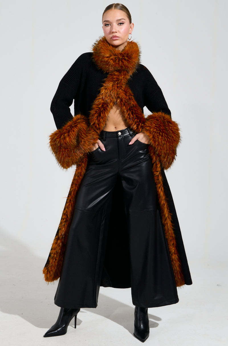 Dressed in a chic winter ensemble, a person stands confidently against a plain backdrop, showcasing the SNOWY NIGHT LONG CARDIGAN IN BLACK with its faux fur collar and cuffs. Their stylish look is completed with wide-leg leather pants and black ankle boots, perfectly capturing the season's fashion-forward style.