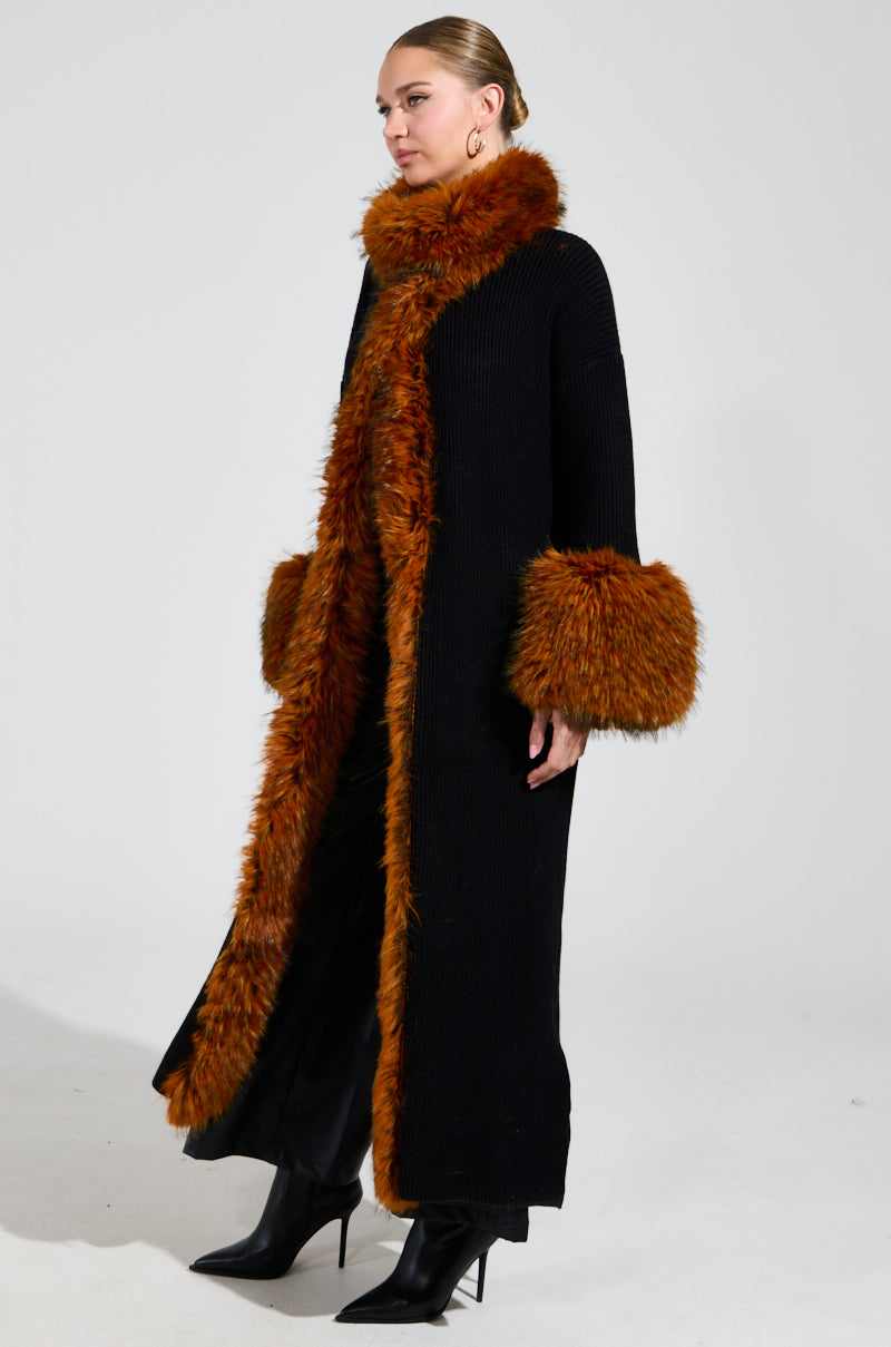 Modeling the SNOWY NIGHT LONG CARDIGAN IN BLACK, a person stands against a gray background. The cardigan features a faux fur collar and brown trim on the cuffs and front edges, perfectly complementing their slicked-back hair and stylish black heeled boots to create a chic winter ensemble.