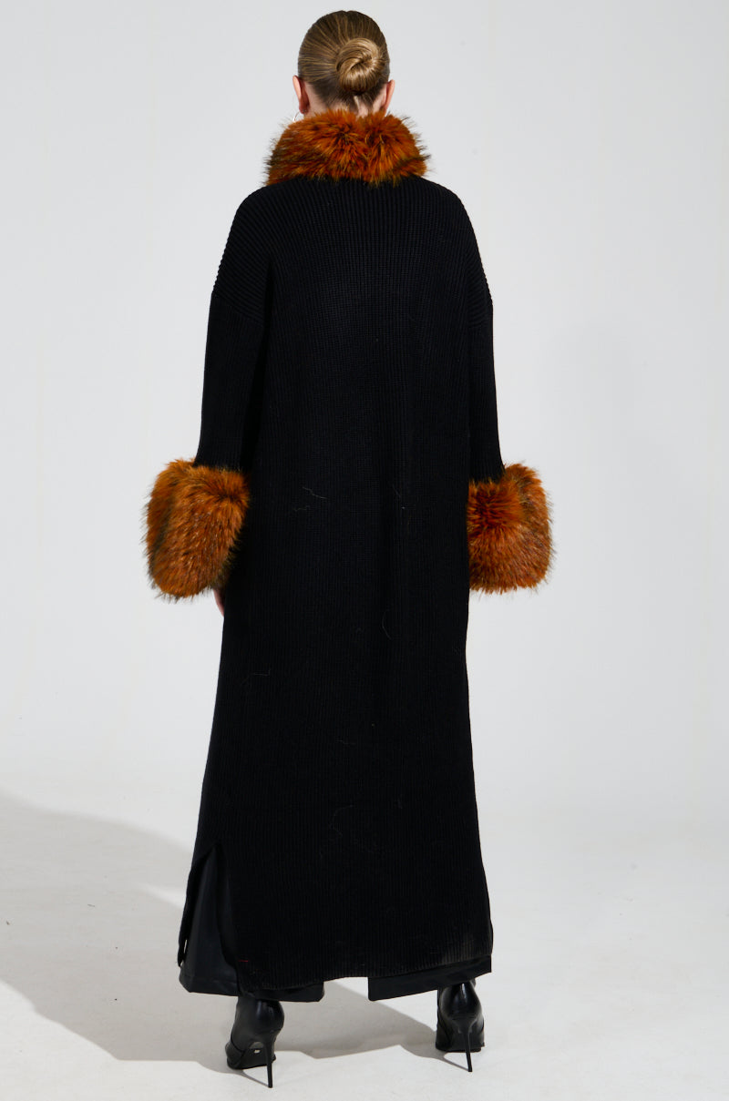 A person is embracing a winter look with the SNOWY NIGHT LONG CARDIGAN IN BLACK, featuring an orange faux fur collar and cuffs. Their hair is styled in a bun as they face away from the camera. The cardigan's hem elegantly reaches their calves, paired with sleek black shoes.