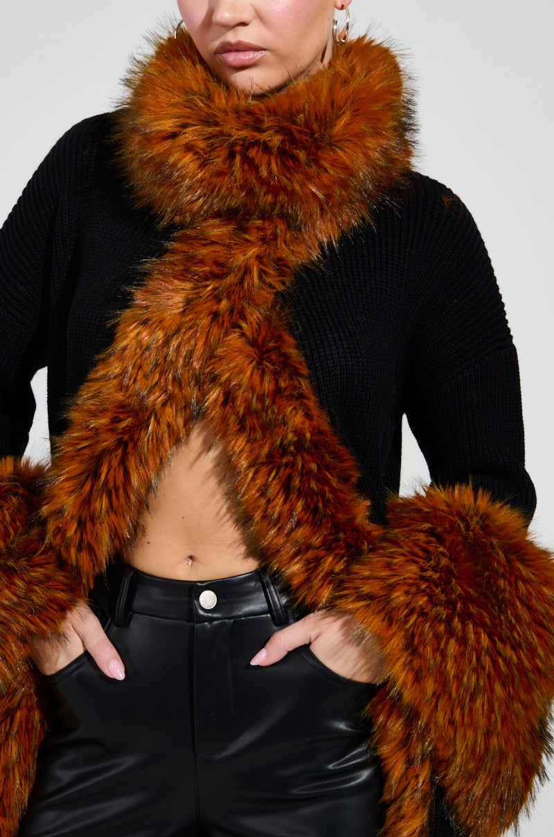 Dressed in the SNOWY NIGHT LONG CARDIGAN IN BLACK, featuring an orange-brown faux fur collar and cuffs, a person stands confidently with hands on hips. The fluffy and vibrant fur contrasts beautifully with sleek leather pants, crafting a chic winter ensemble that showcases stylish fashion details.