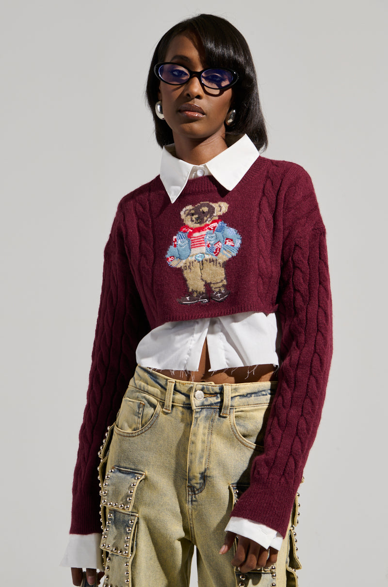 A stylish individual wears the AKIRA Label TEDDY SWEATER, showcasing an embroidered bear design over a crisp white collared shirt. Complemented by light brown pants with decorative details, they flaunt long, dark hair accented with oval-shaped sunglasses and elegant earrings.