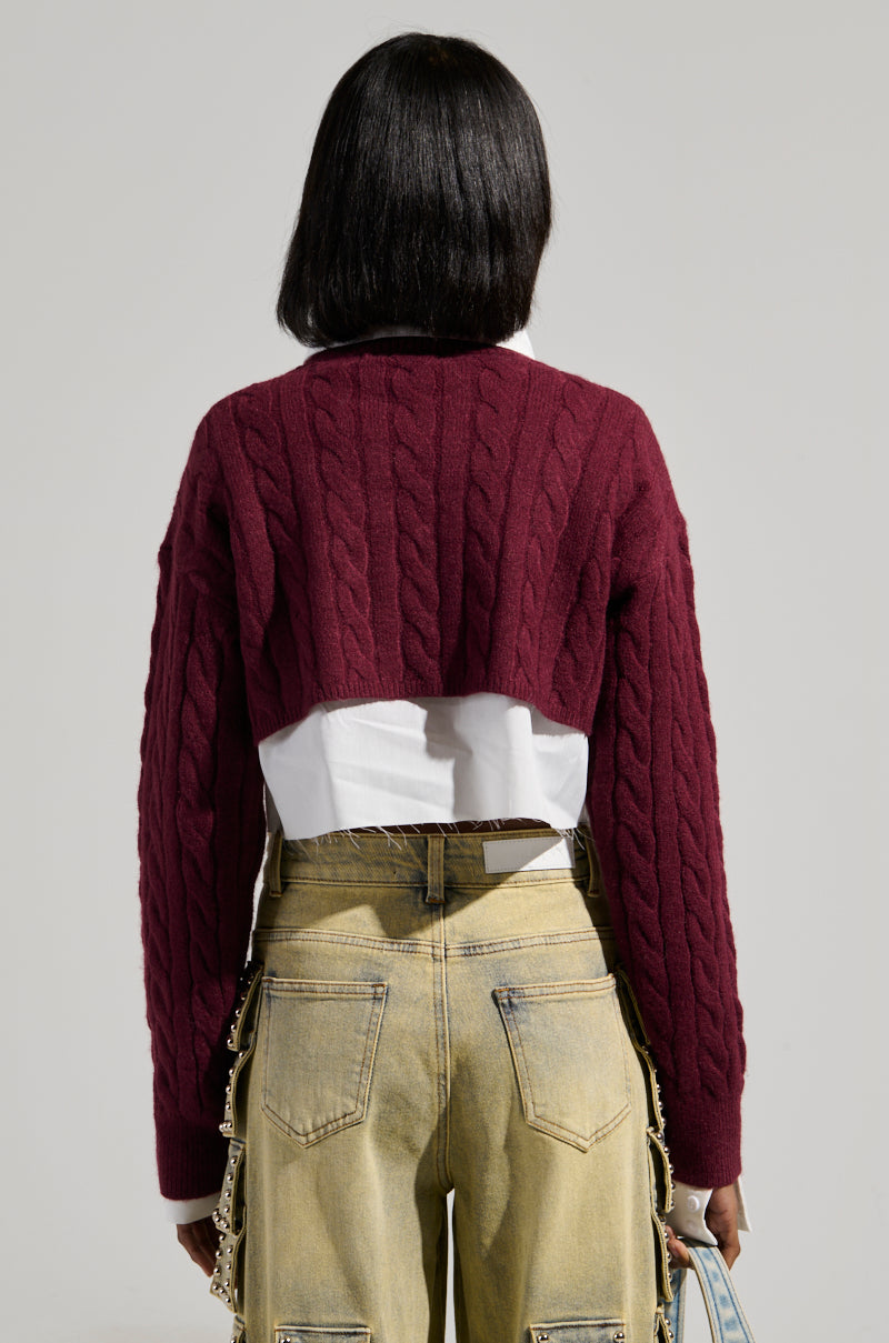 A woman with shoulder-length dark hair is seen from behind, wearing a maroon TEDDY SWEATER by AKIRA Label over a white shirt. She has on beige pants with chain details and holds a light blue handbag. The background is a plain light gray.