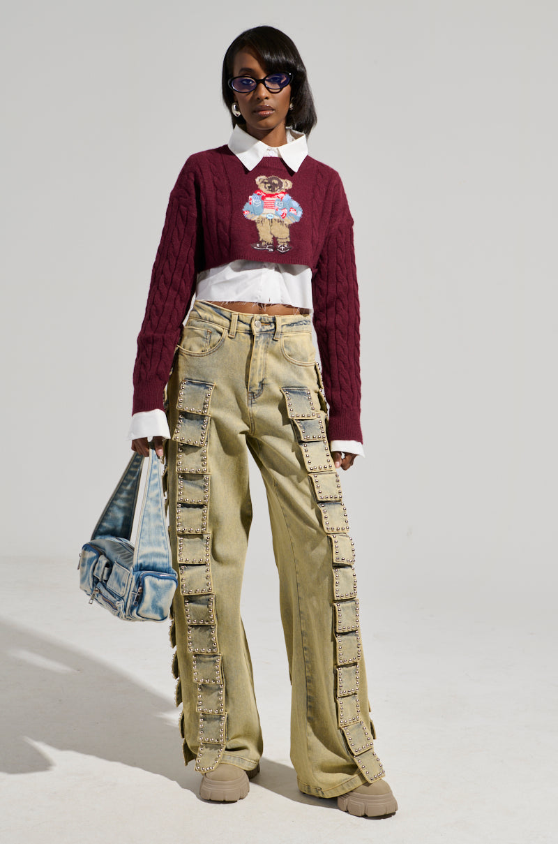 A person is dressed in a burgundy TEDDY SWEATER from the AKIRA Label, featuring a graphic design. Underneath is a white collared shirt, paired with unique tan pants that boast stitched rectangular patterns. They're accessorizing with sunglasses and carrying a denim bag while standing against a plain background.