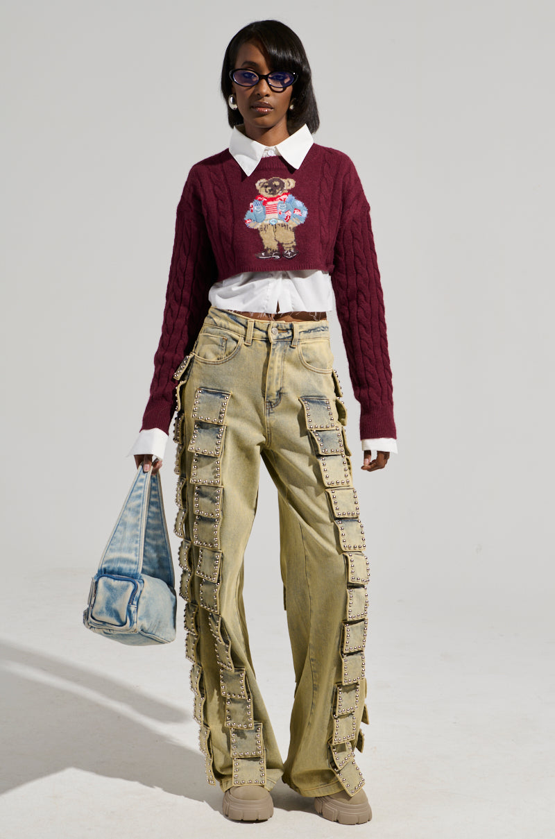 A person wearing the TEDDY SWEATER from AKIRA Label, which showcases an embroidered bear graphic, over a white shirt, paired with wide-legged khaki pants featuring buckle details. They complete their look with sunglasses and a denim handbag, effortlessly combining casual and quirky fashion elements.