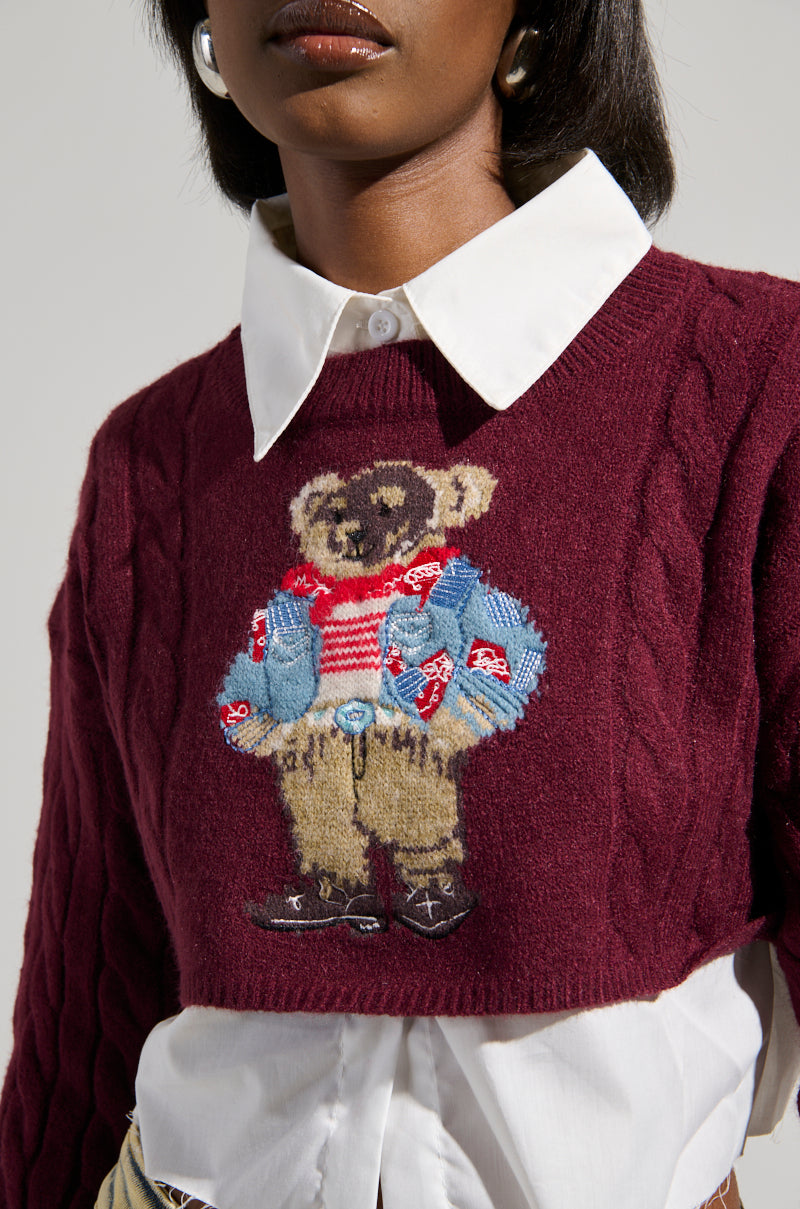 A person wearing the TEDDY SWEATER from the AKIRA Label, showcasing a burgundy cable-knit design adorned with an embroidered bear graphic layered over a white collared shirt. The delightful bear is dressed in a red striped shirt, blue jacket, and tan pants set against a simple, neutral backdrop.