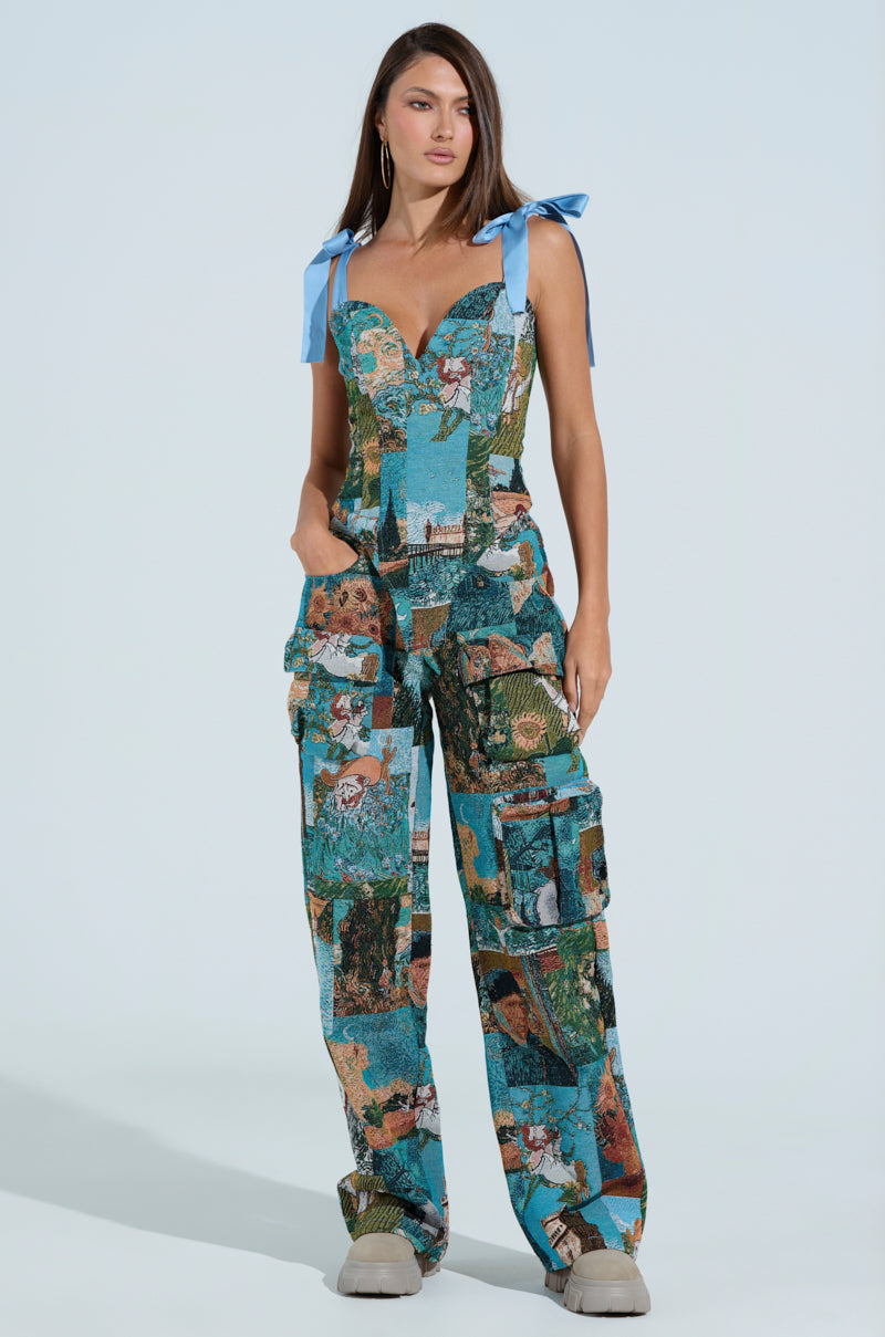 A woman confidently models the LOCKED AND LOADED TROUSER, featuring artistic Impressionist-inspired patterns and blue bow straps. With its vibrant design and multiple pockets, she poses against a plain, light background.