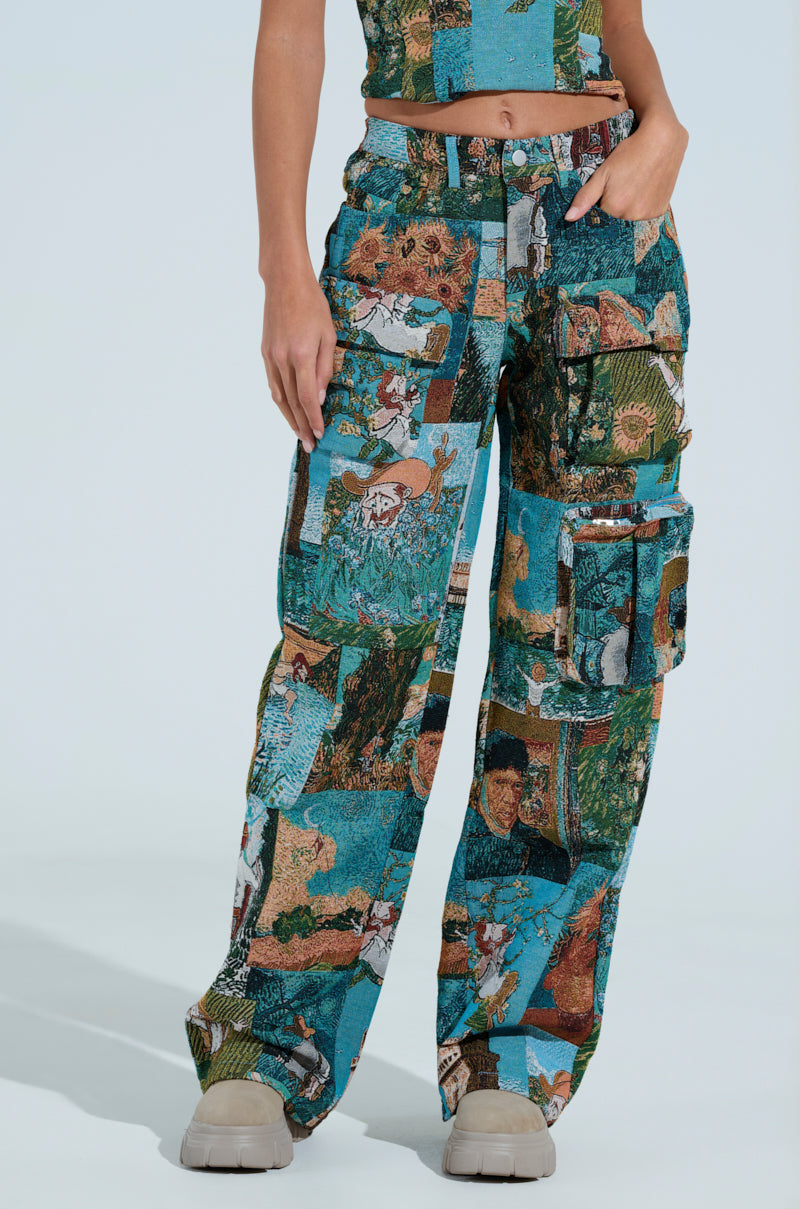 A person wears a vibrant two-piece featuring artistic collage patterns in blues, greens, and browns. The high-rise LOCKED AND LOADED TROUSER includes multiple pockets and the look is finished with chunky beige boots.