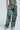 A person wears a vibrant two-piece featuring artistic collage patterns in blues, greens, and browns. The high-rise LOCKED AND LOADED TROUSER includes multiple pockets and the look is finished with chunky beige boots.