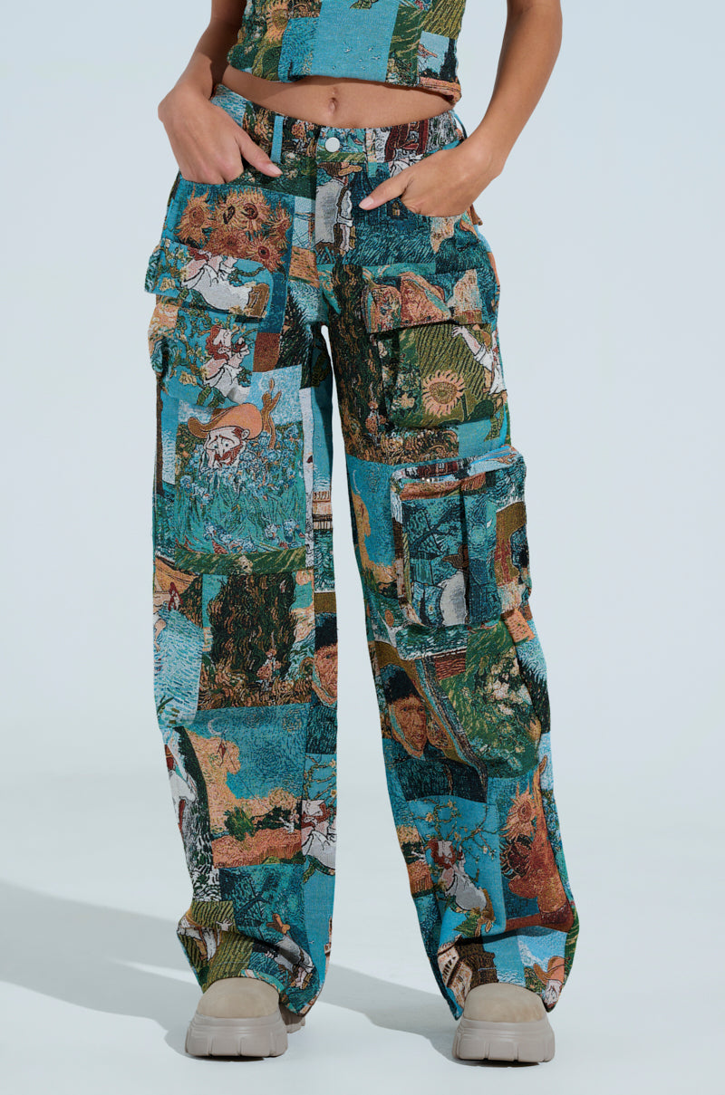 A person poses against a light blue backdrop in the LOCKED AND LOADED TROUSER, featuring vibrant patchwork cargo pants with a high-rise fit. The colorful collage pattern in shades of blue, green, and brown complements the matching top and is stylishly paired with beige shoes.