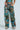 A person poses against a light blue backdrop in the LOCKED AND LOADED TROUSER, featuring vibrant patchwork cargo pants with a high-rise fit. The colorful collage pattern in shades of blue, green, and brown complements the matching top and is stylishly paired with beige shoes.