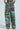 Wearing vibrant LOCKED AND LOADED TROUSERs, featuring artistic landscapes and figures in a colorful patchwork against a light background, one showcases an ensemble complemented by a matching top that radiates impressionist-inspired artwork flair.