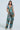 A person in a LOCKED AND LOADED TROUSER with a colorful collage pattern and blue bow straps stands against a light background. Reminiscent of impressionist artwork, it features various prints. They have long hair, hoop earrings, and beige shoes.