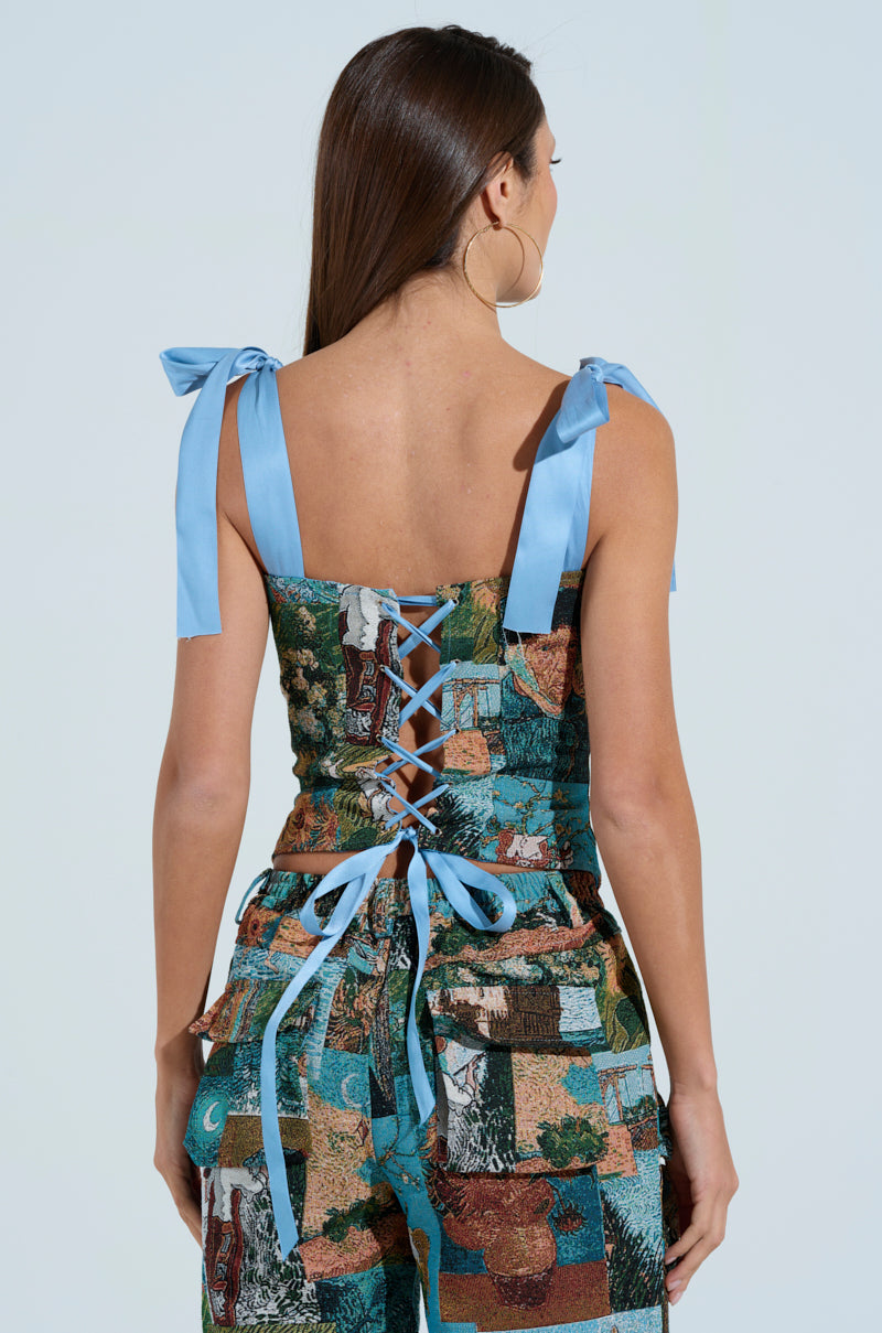 A woman with long brown hair, seen from the back, wears the LOCKED AND LOADED CORSET—a colorful jacquard two-piece with a corset-style lace-up back and blue ribbon shoulder ties, featuring intricate green, blue, and brown motifs.