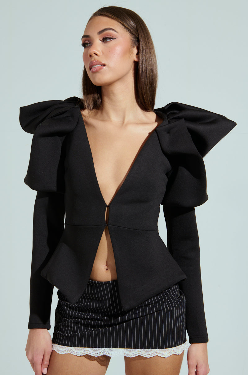A woman pairs the MILA FITTED BLOUSE IN BLACK, featuring a plunging neckline and large shoulder bows, with a black pinstripe skirt with a lace hem, set against a light blue backdrop that enhances the chic contrast of her outfit.