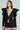 A woman pairs the MILA FITTED BLOUSE IN BLACK, featuring a plunging neckline and large shoulder bows, with a black pinstripe skirt with a lace hem, set against a light blue backdrop that enhances the chic contrast of her outfit.