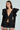 A woman in a chic outfit poses against a light background, wearing the black Mila Fitted Blouse with large shoulder bows and a plunging V-neckline, paired with a pinstripe mini skirt featuring a lace hem. Her straight, long hair completes her stylish appearance.