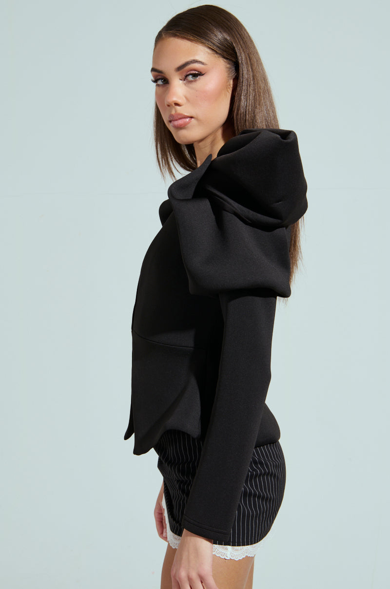 A woman with long brown hair poses in a black MILA FITTED BLOUSE featuring a peplum silhouette and oversized shoulders, paired with pinstripe shorts. She stands against a light background, gazing over her shoulder.