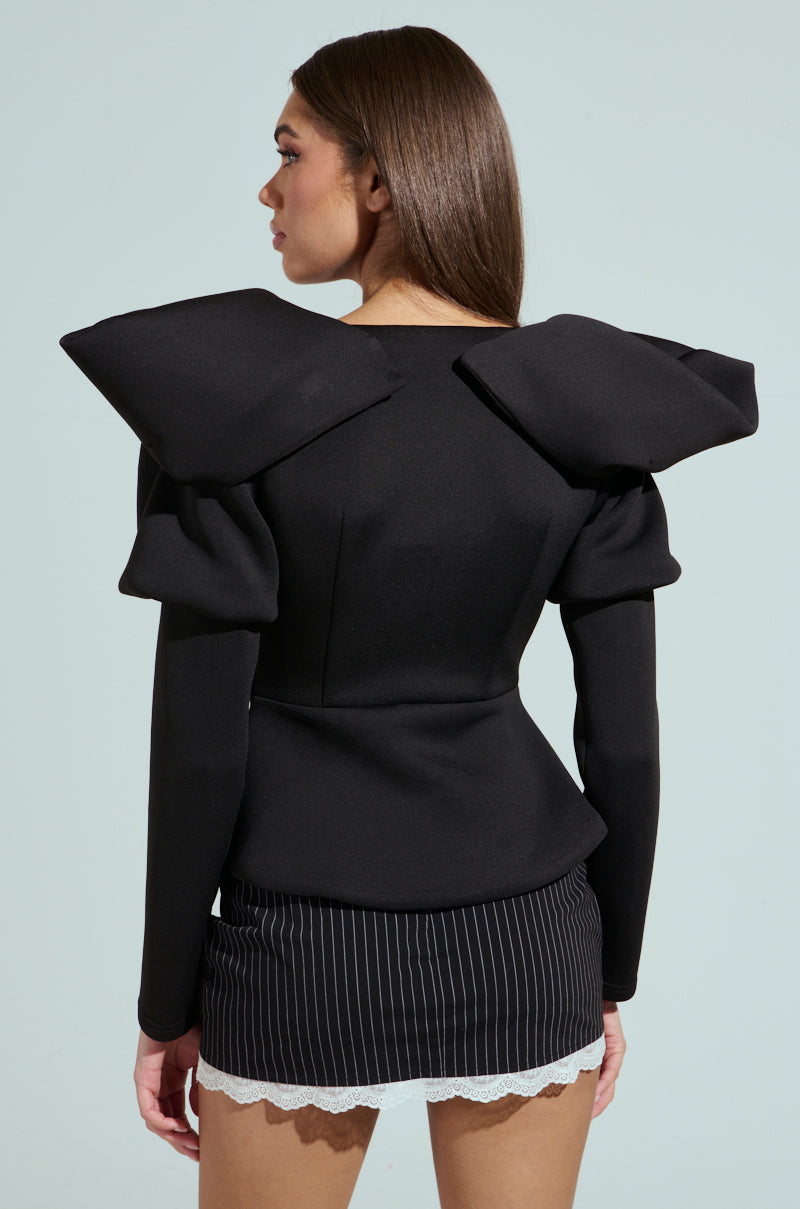 A person with long brown hair wears a distinctive Mila Fitted Blouse in black featuring large, angular shoulder pads and a plunging neckline, paired with a pinstriped skirt adorned with white lace trim against a soft green background.