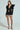 A woman poses against a plain backdrop wearing the MILA FITTED BLOUSE IN BLACK, featuring a plunging neckline and large shoulder ruffles, paired with a short pinstriped skirt with a white lace hem and strappy black high heels.