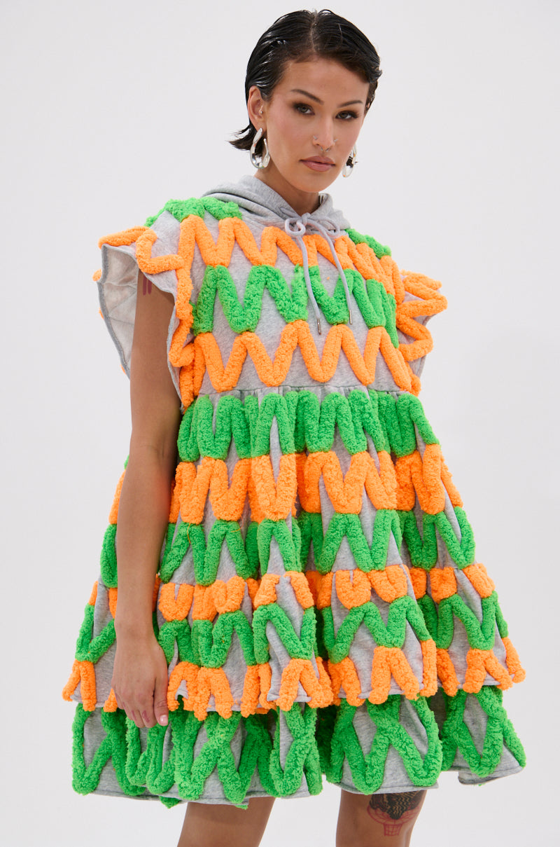 A woman poses against a white background wearing the LOLO ZIGZAG MINI DRESS, a loose, short-sleeved mini dress with green and orange textured zigzag appliqués and ruffled layers, styled with silver earrings.