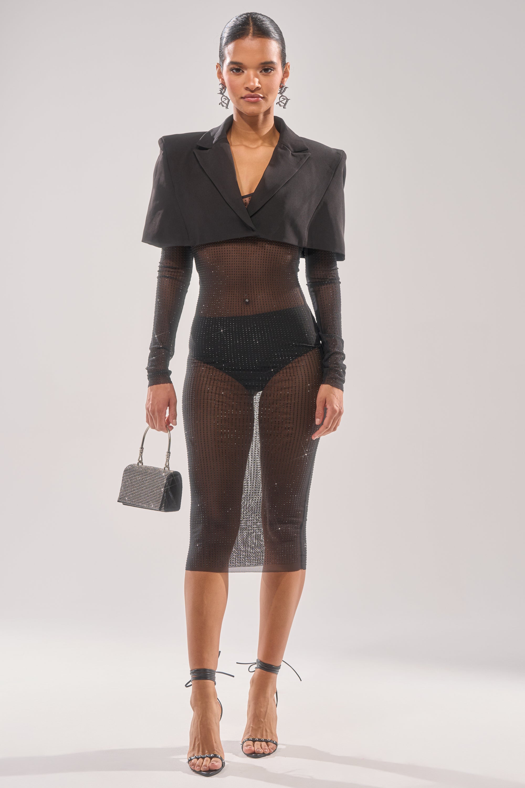 A woman models the SHE MEANS BUSINESS RHINESTONE MIDI DRESS—sheer and black with sparkly rhinestones—over black undergarments, styled with a cropped blazer, strappy heels, statement earrings, and a small handbag against a light background.