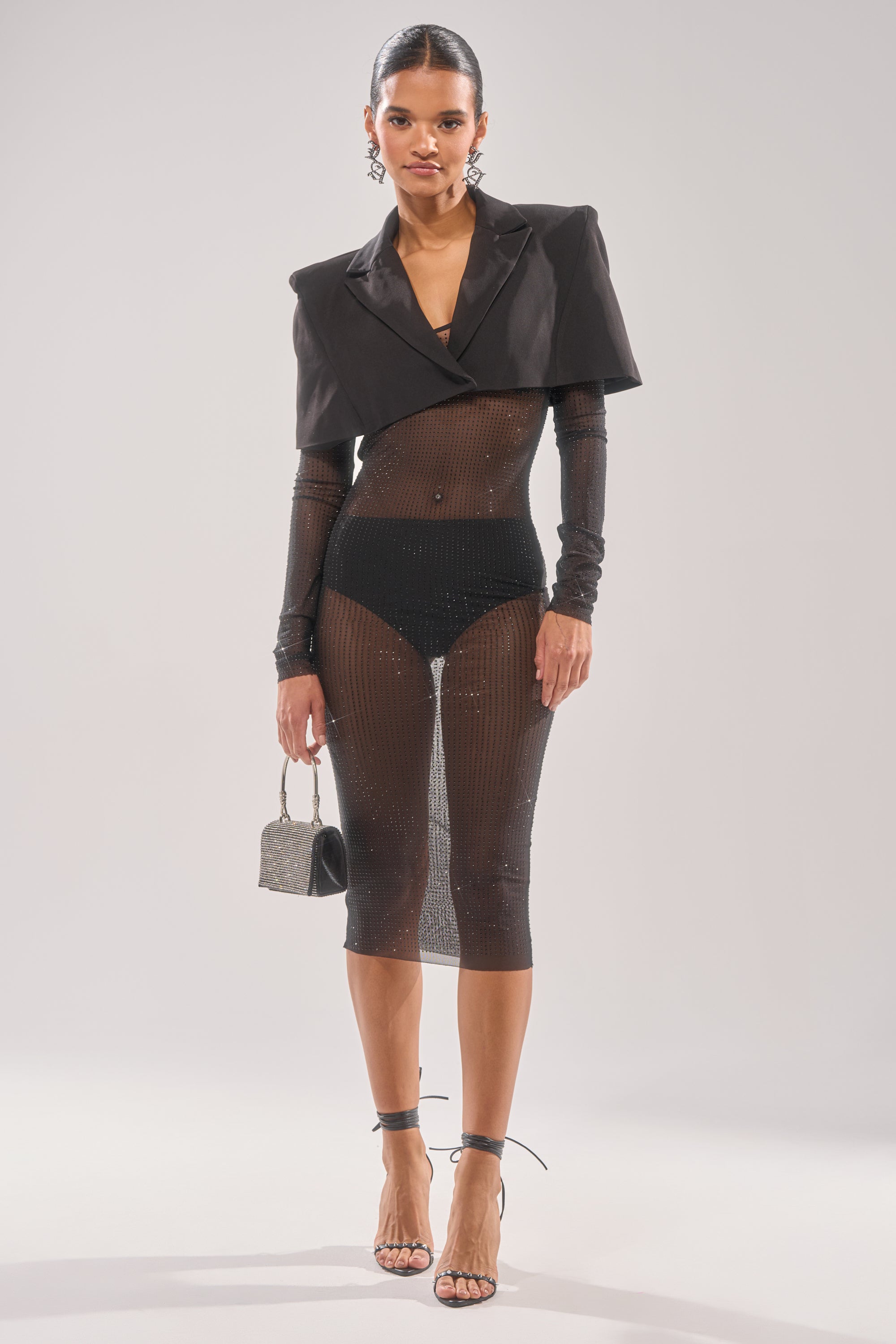 A woman poses in the SHE MEANS BUSINESS RHINESTONE MIDI DRESS, featuring sheer black fabric, rhinestones, three-quarter sleeves, and black high-waisted underwear. She completes the look with a cropped blazer, heels, earrings, and a metallic handbag.