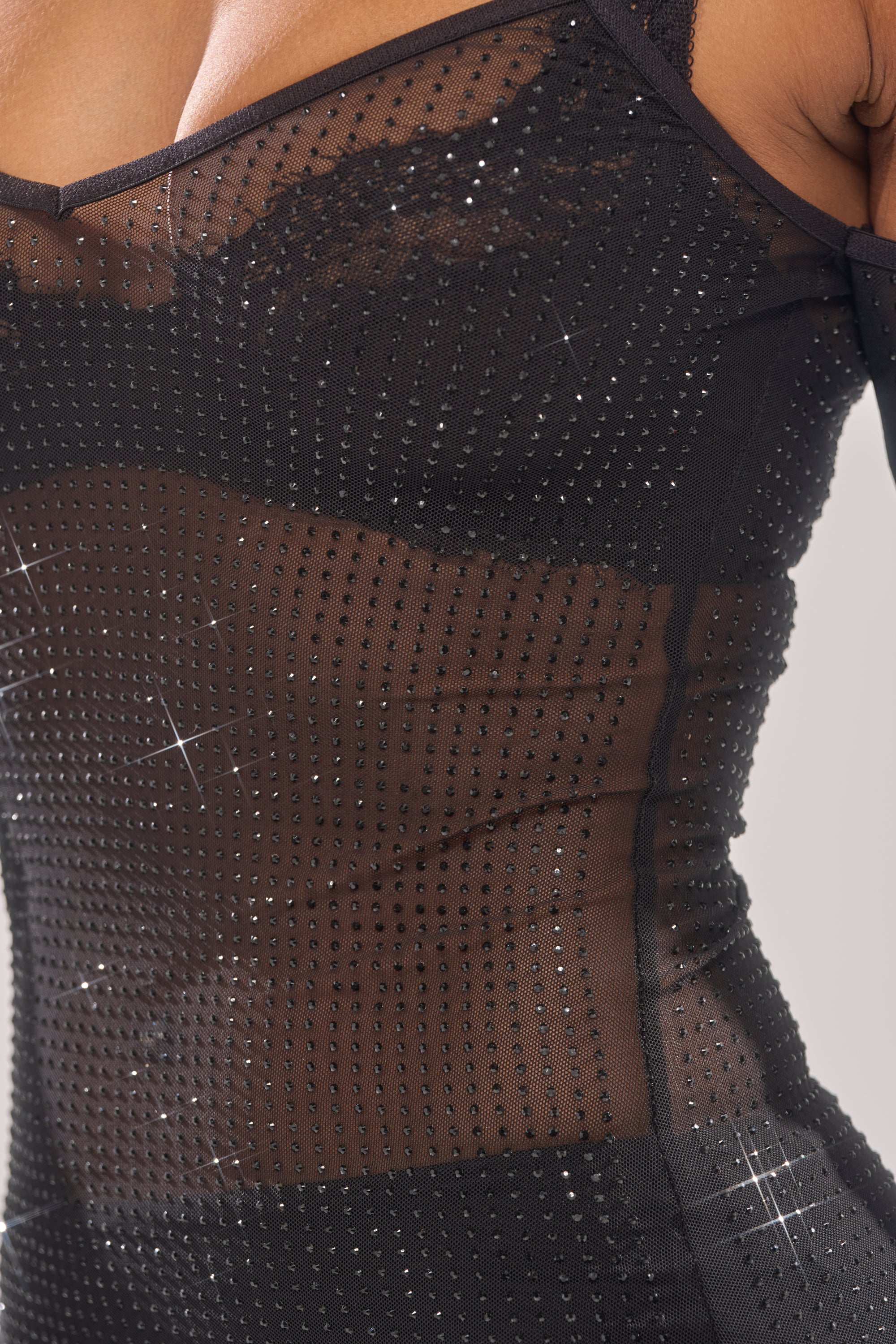A close-up of a person wearing the SHE MEANS BUSINESS RHINESTONE MIDI DRESS—a sheer black dress with thin straps and sparkling rhinestone details, revealing some skin beneath.