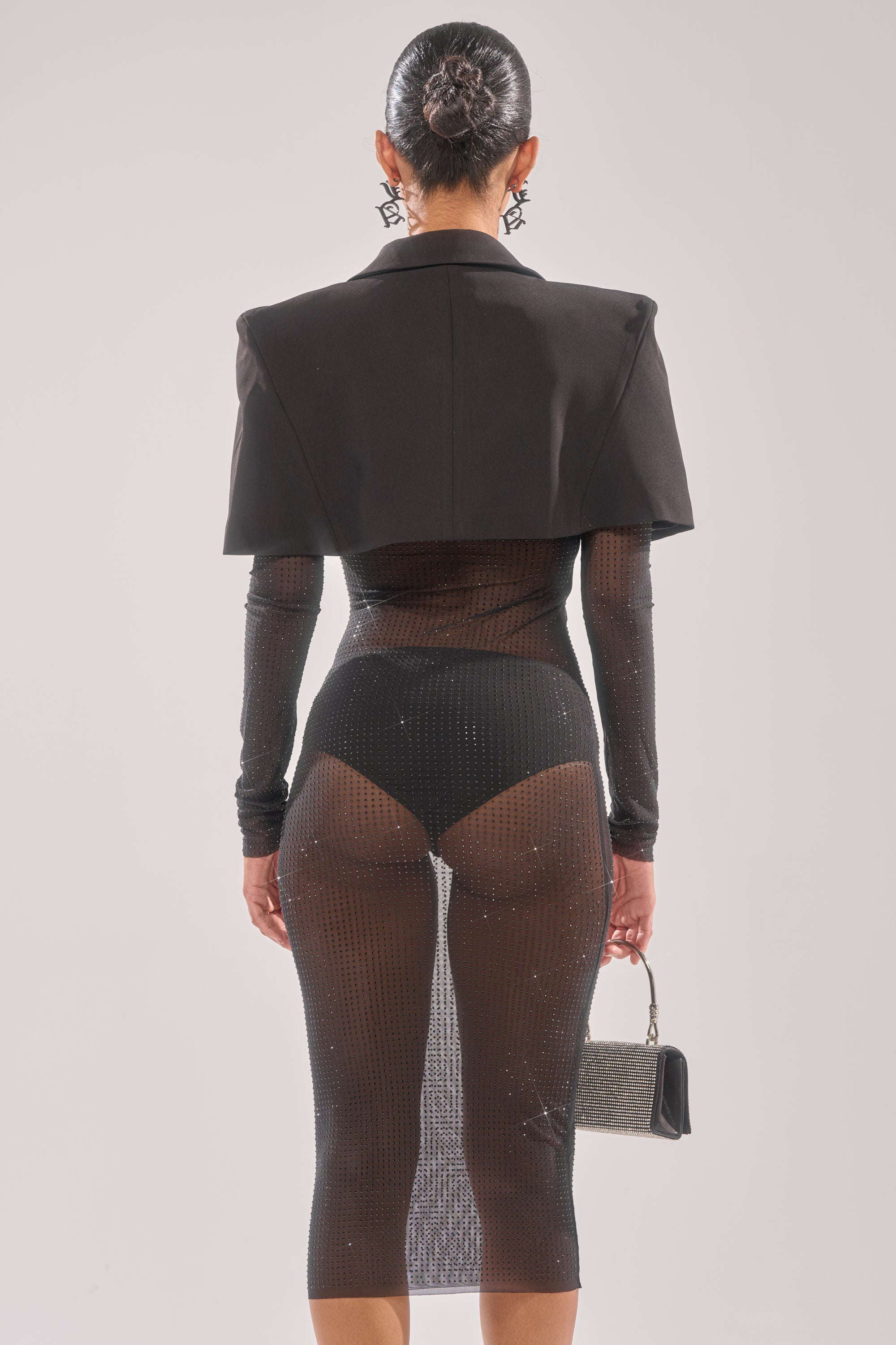 Wearing the SHE MEANS BUSINESS RHINESTONE MIDI DRESS with visible black undergarments and a cropped jacket, a person stands facing away, holding a small handbag and sporting a sleek bun hairstyle.