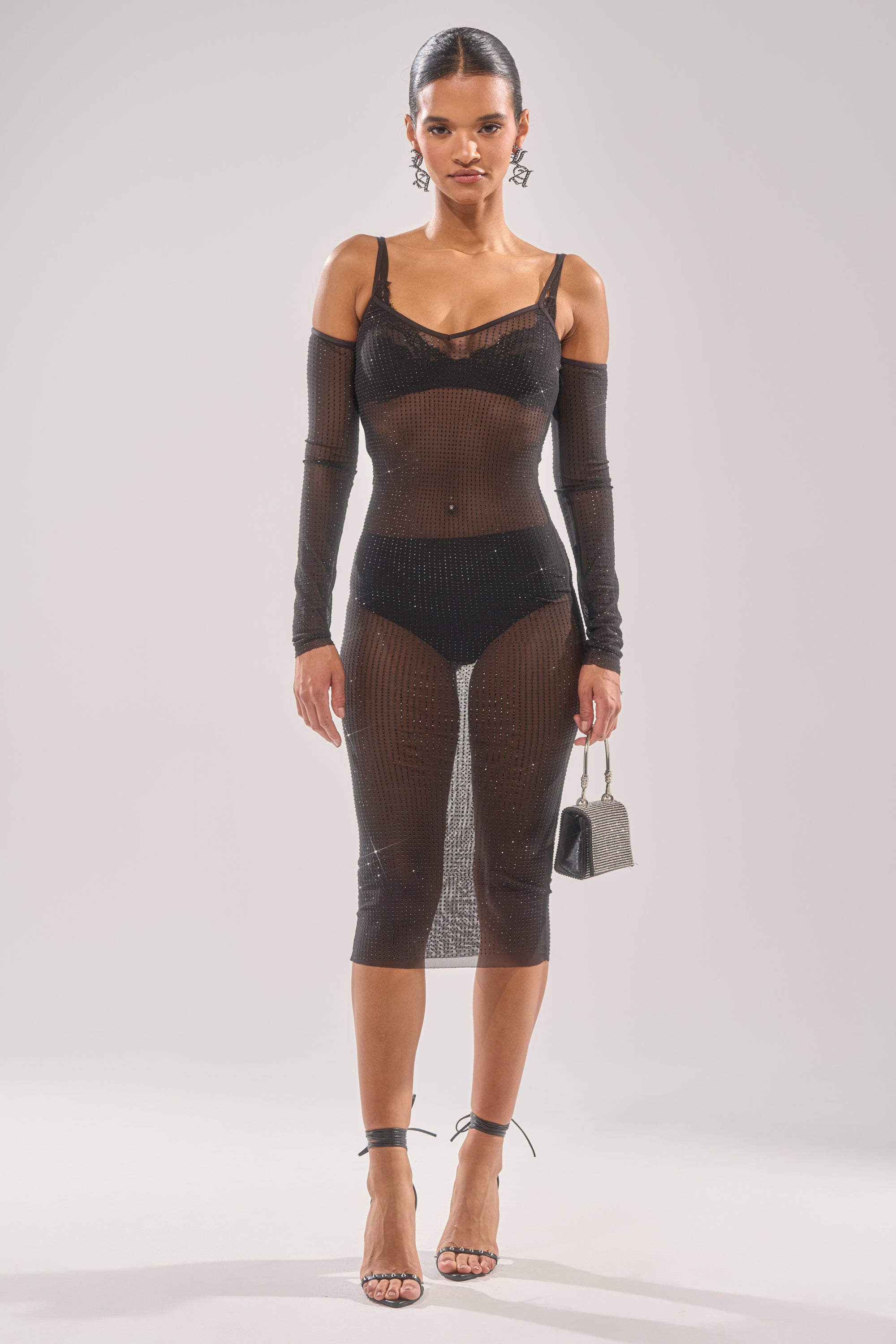 She faces forward in the SHE MEANS BUSINESS RHINESTONE MIDI DRESS—a sheer black cold-shoulder style with long sleeves, worn over black undergarments. She accessorizes with a silver handbag, high-heeled sandals, and slicked-back hair.