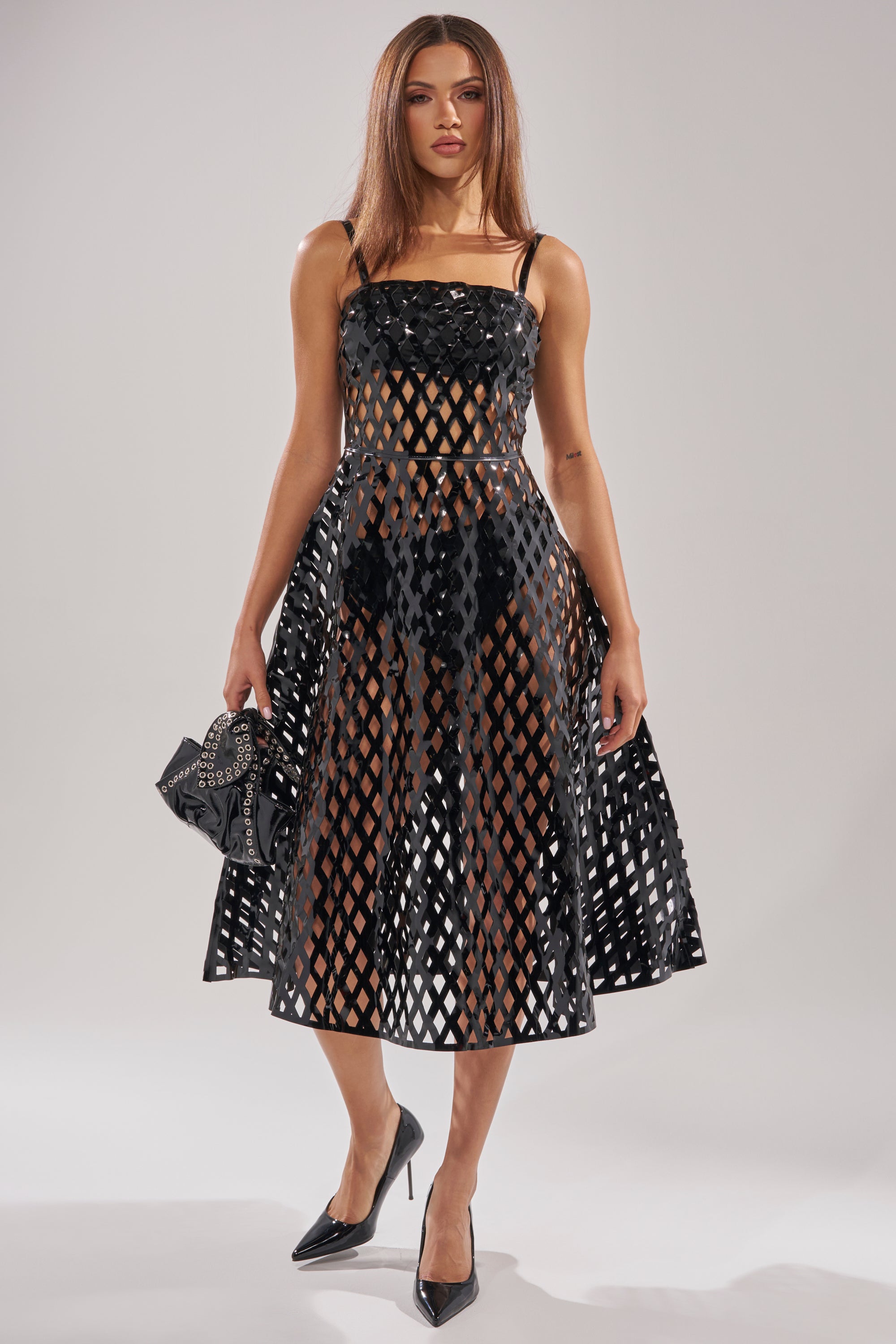 A woman in the SEXY AS CAN BE MIDI DRESS—a sleeveless, black cut-out midi with a lattice-patterned sheer look—pairs it with black heels and a studded clutch, standing against a plain light background.