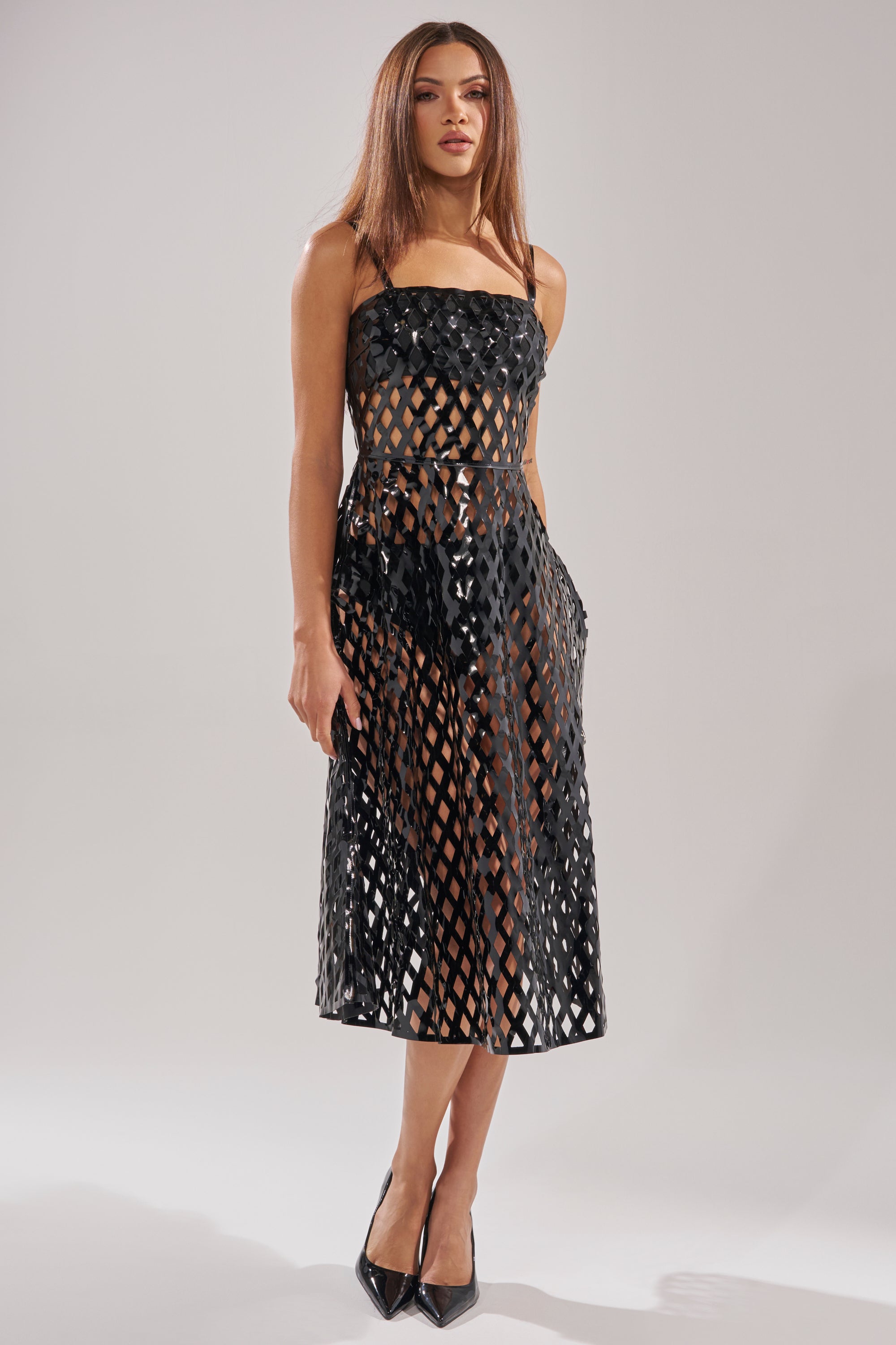 A woman poses against a plain background wearing the SEXY AS CAN BE MIDI DRESS— a sleeveless black cut-out design with a lattice pattern, sheer skirt, and black undergarments. She has straight brown hair and black pointed-toe heels.