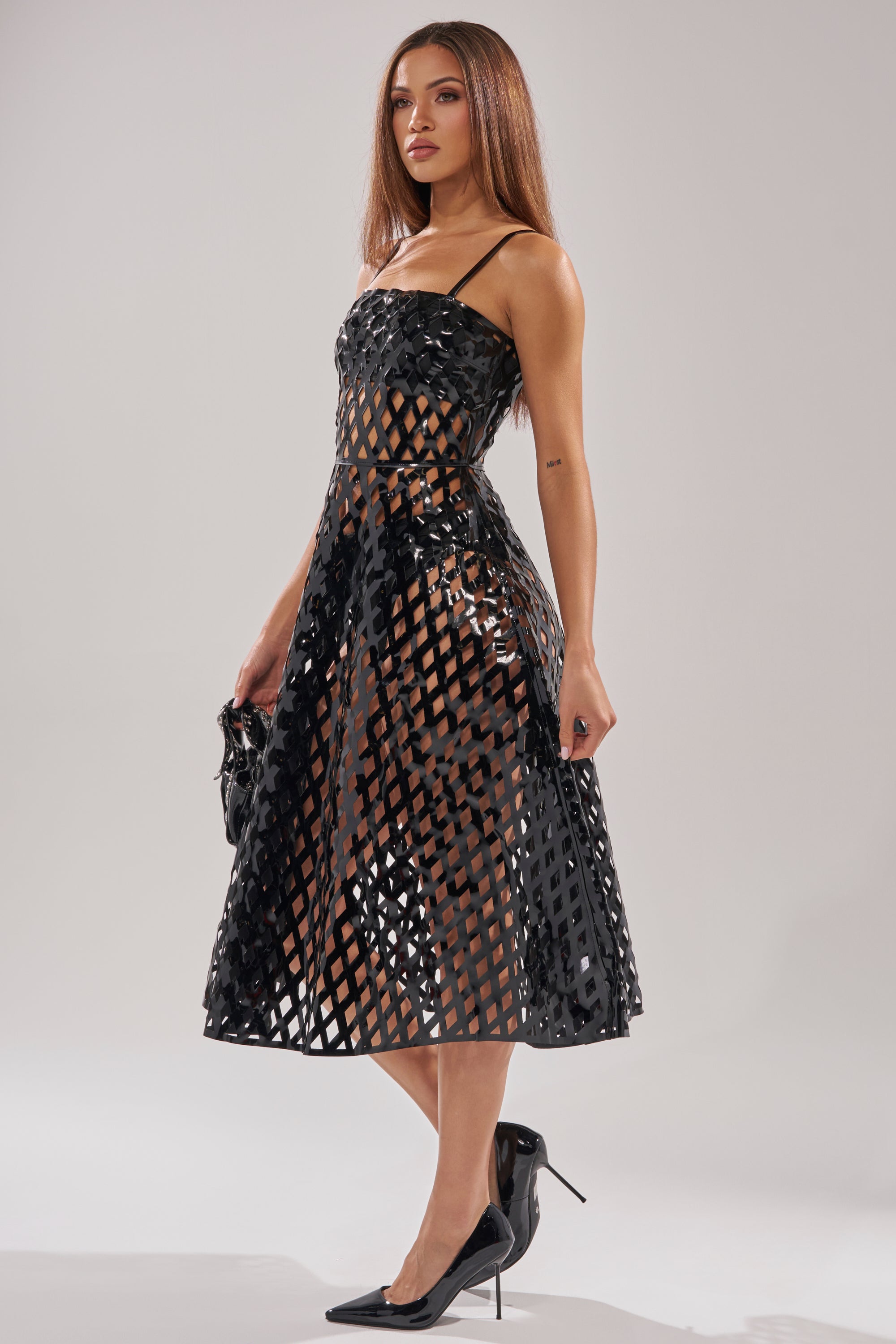 A woman wears the SEXY AS CAN BE MIDI DRESS, featuring a black lattice pattern with semi-transparent diamond cutouts and a flared skirt. She completes the look with a black clutch and pointed high heels.