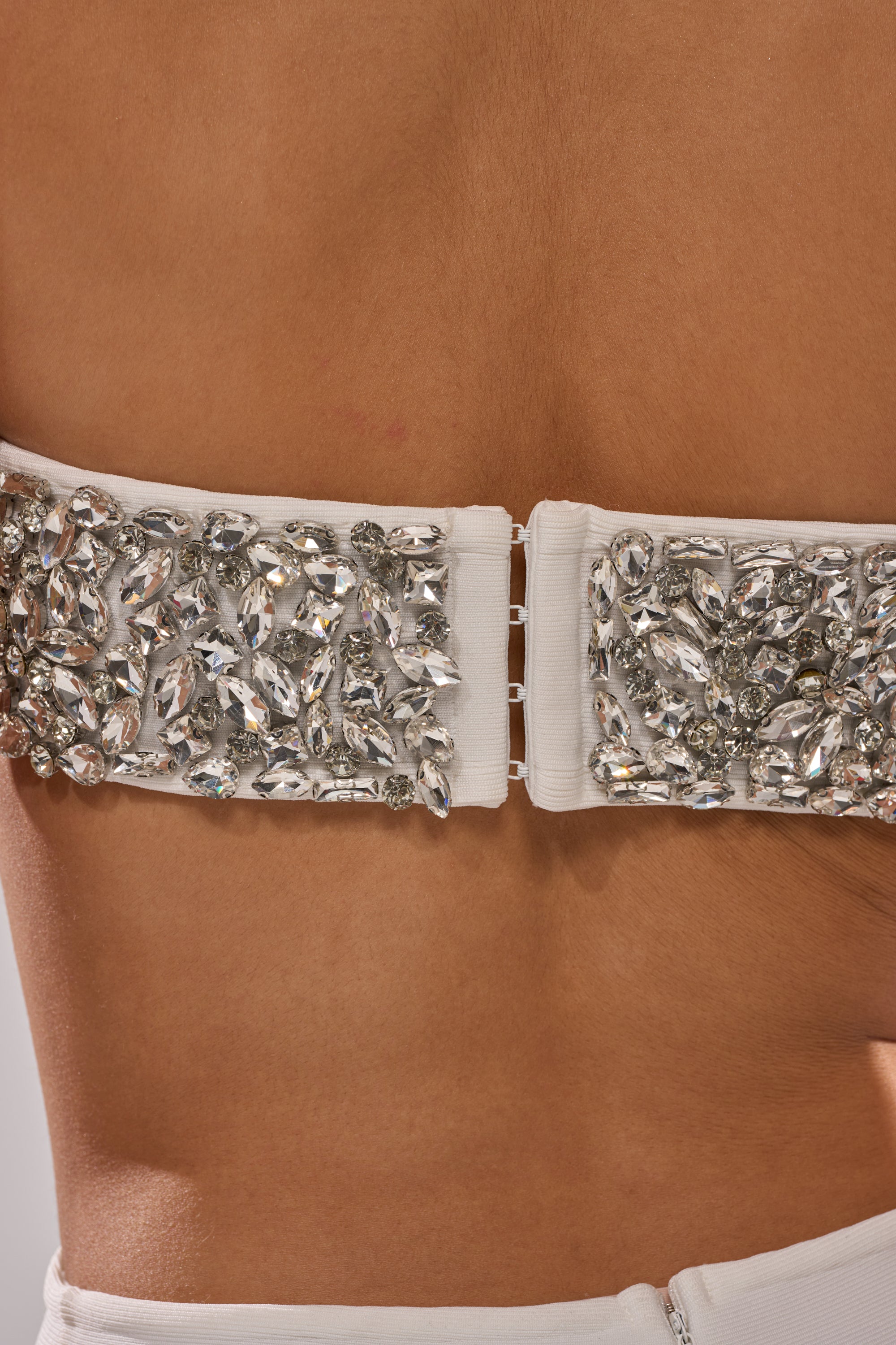 Close-up of a tan back wearing the RICH AF RHINESTONE CUTOUT MINI DRESS, a white bandage dress with a jeweled strap adorned with sparkling rhinestones in various shapes and a hook-and-eye closure for an elegant, decorative look.