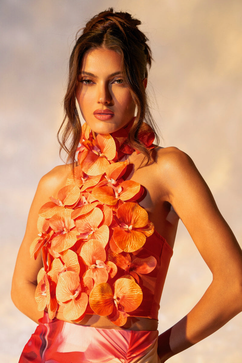 A woman poses in the FLOWER BOMB TOP, featuring a mock neck and bright orange floral appliqués. Her hair is styled up with loose strands, set against a softly blurred background with warm lighting.
