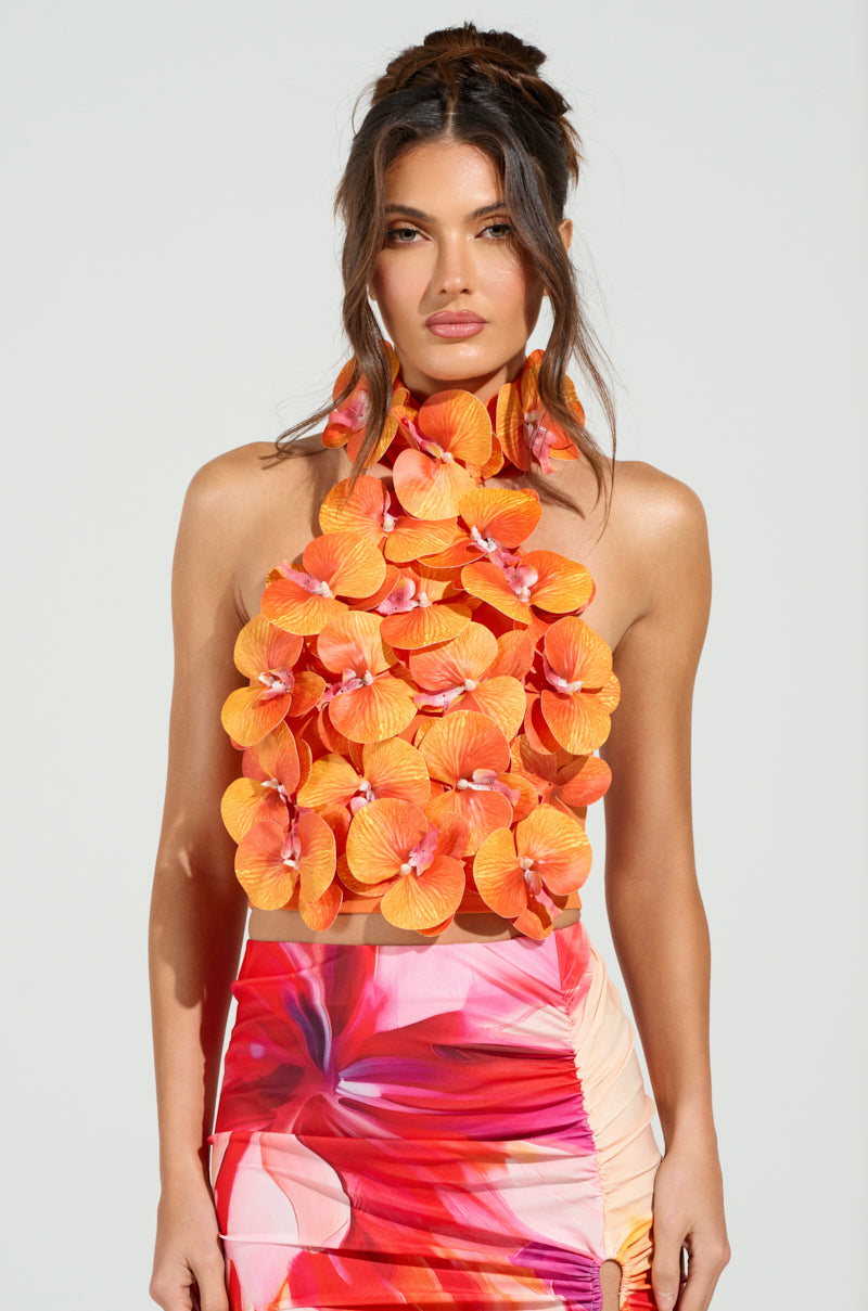 A woman with long brown hair in an updo wears the FLOWER BOMB TOP with orange floral appliqués, paired with a colorful skirt in pink, red, and white hues. The cropped ensemble stands out against a plain light background.