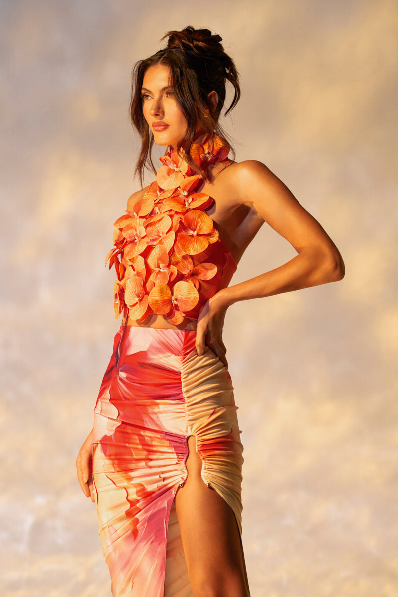 A woman wears the FLOWER BOMB TOP with tropical floral appliqués on the bodice. The vibrant top in pink and orange hues complements her elegant updo as she poses against a softly blurred background.