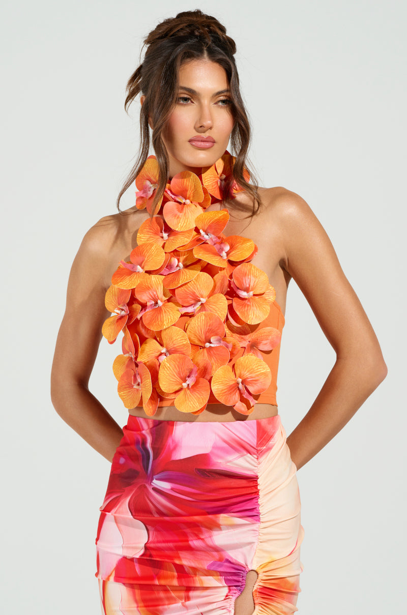 A woman in the FLOWER BOMB TOP, decorated with bold orange flowers, complements her look with a vibrant pink and orange skirt featuring tropical floral appliqués. Her hair is styled in an updo against a plain backdrop.