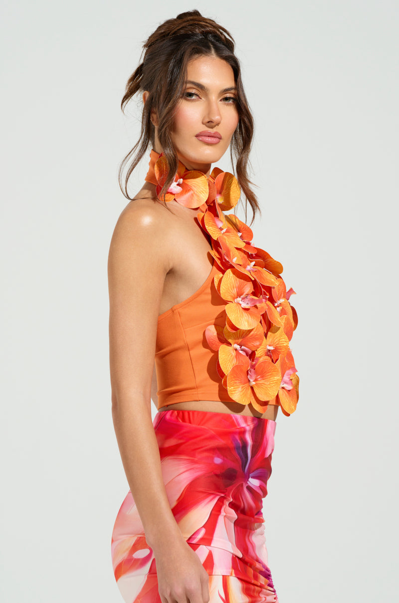A woman poses confidently in the FLOWER BOMB TOP. The mock neck top features tropical floral appliqués, paired with a pink, red, and white skirt. Her hair is styled in loose waves with part pulled back against a neutral background.
