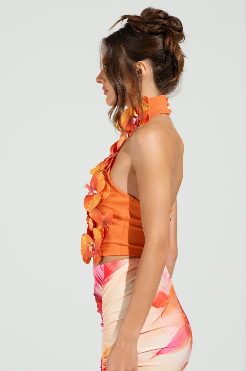A woman with brown hair styled in an updo wears the vibrant orange FLOWER BOMB TOP with tropical floral appliqués, and her skirt features shades of pink and orange. She looks to the right against a plain background.