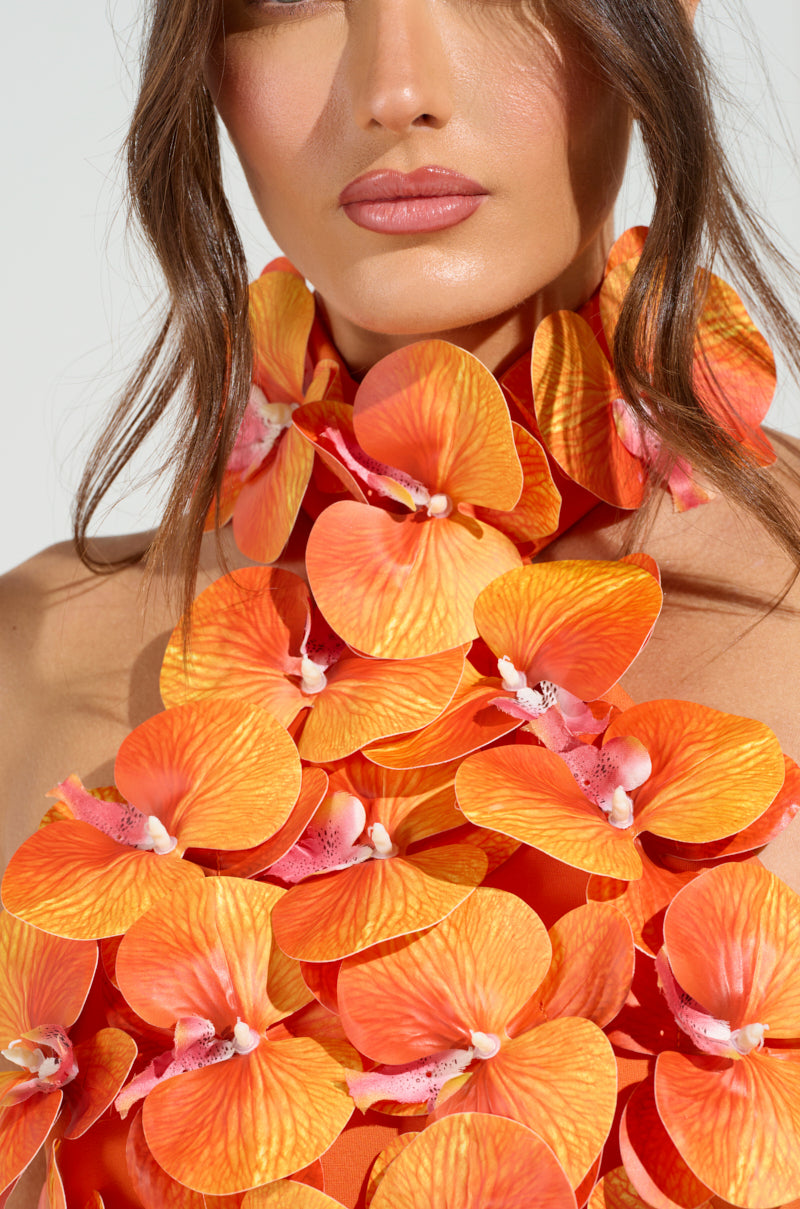 A woman with long brown hair models the FLOWER BOMB TOP, featuring a vibrant orange orchid arrangement around her neck and shoulders. The vivid petals contrast beautifully with her neutral makeup and the light background, reminiscent of tropical floral appliqués.