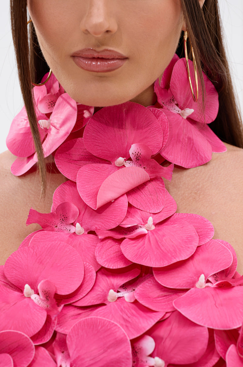 A woman with straight brown hair and large gold hoop earrings wears the FLOWER BOMB TOP IN FUCHSIA, featuring artificial orchid petals that cover her chest and neck. The background is plain white. 9