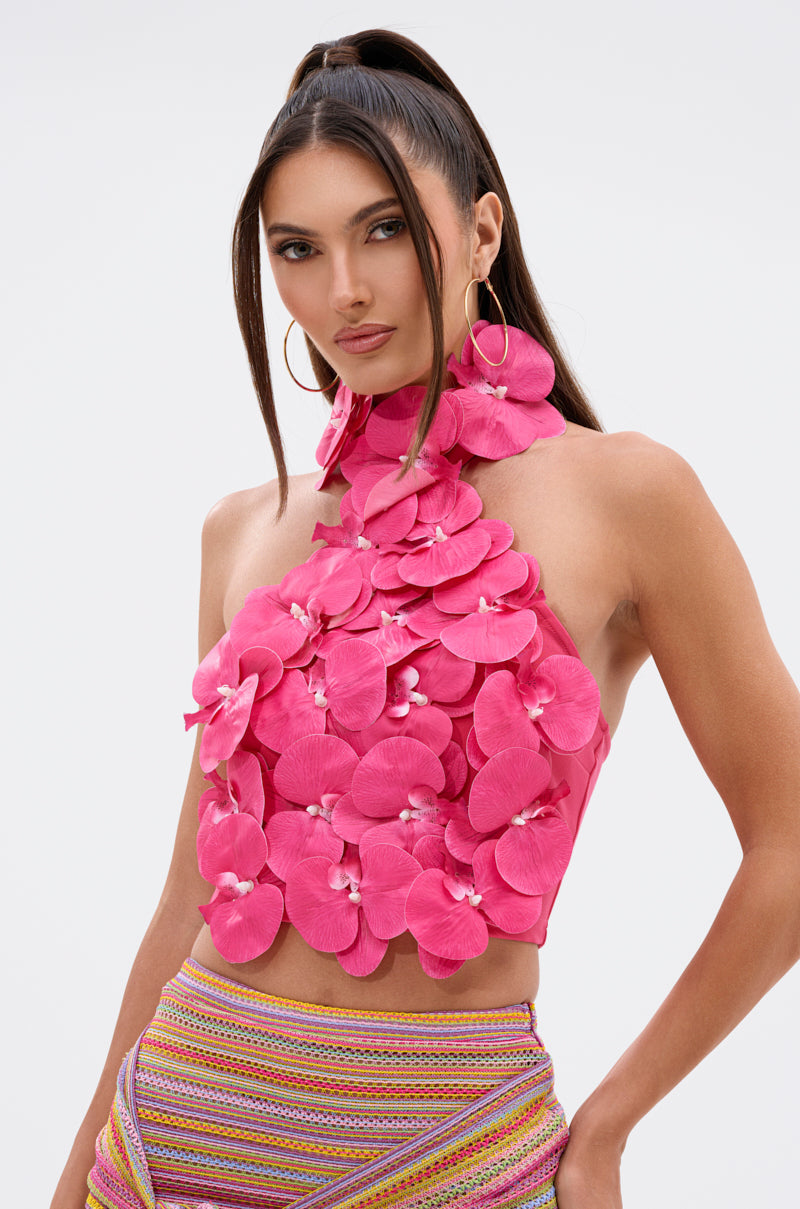 A woman with long brown hair in a high ponytail wears the FLOWER BOMB TOP IN FUCHSIA, featuring bright pink 3D orchid appliqués, and colorful striped pants. She looks confidently at the camera against a plain background. 1
