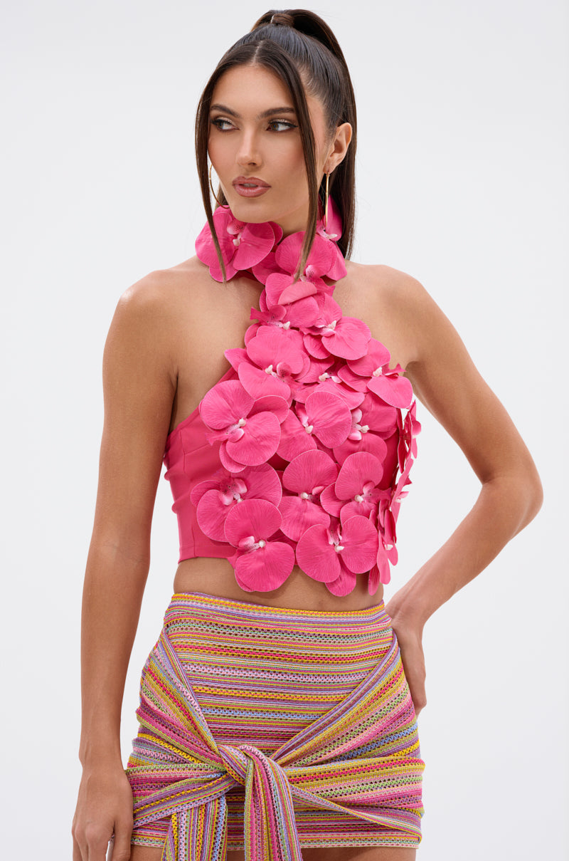 A woman models the FLOWER BOMB TOP IN FUCHSIA, a halter with 3D orchid appliqué petals, styled with a multicolored striped skirt with a knot detail, set against a plain white background. 2