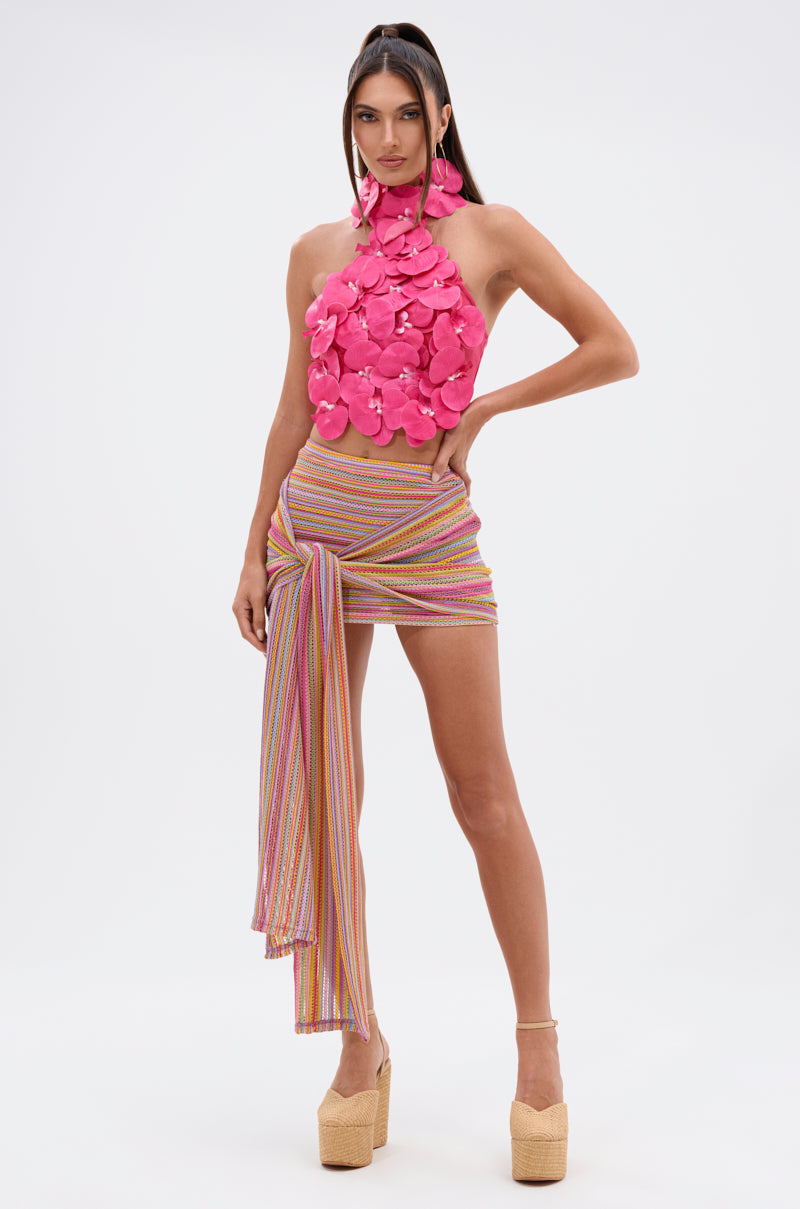 A woman poses confidently in the FLOWER BOMB TOP IN FUCHSIA—a sleeveless halter top with 3D orchid appliqué—paired with a multicolored striped mini skirt and platform heels against a plain white background. 7