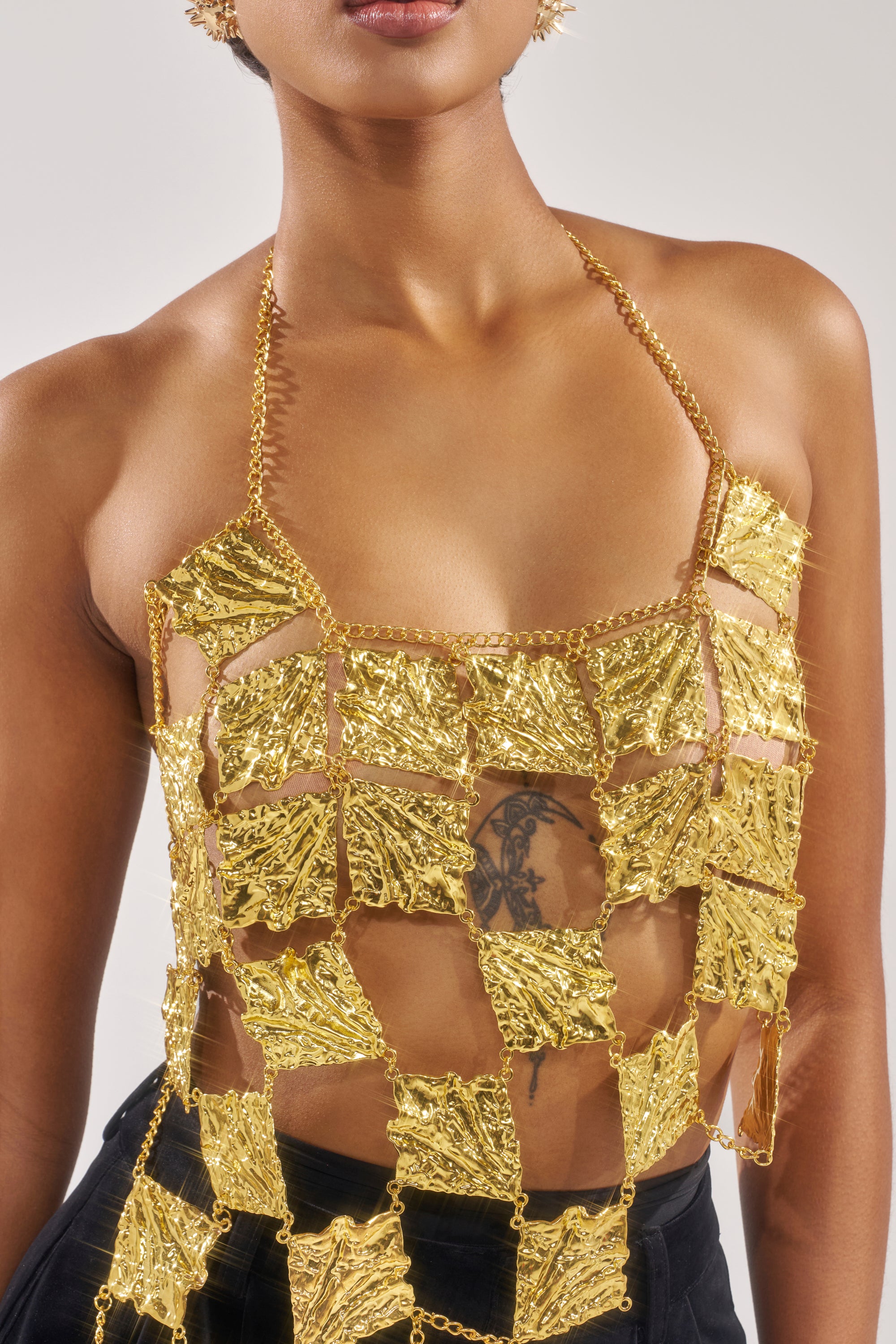 A woman wears the SHOW ME WHAT YOU GOT METAL CHAIN TOP—a sculptural gold piece with textured squares linked by chains, revealing her midriff and a chest tattoo. She finishes the look with matching gold earrings. 7