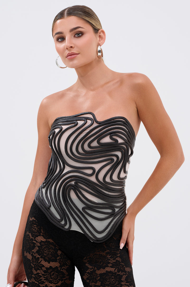A woman poses confidently in the CRAZY IN LOVE CORSET—strapless mesh with black wavy sculptural lines—paired with black lace pants, hoop earrings, and slicked-back hair against a light background.