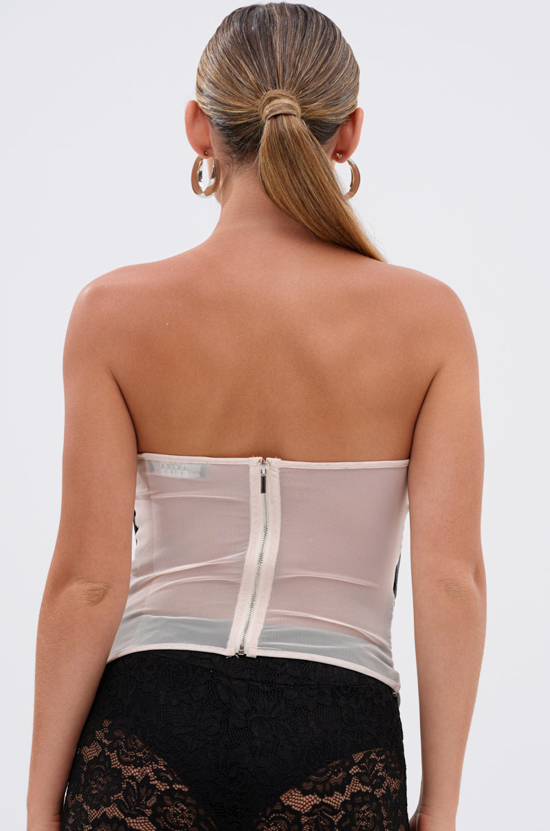 A woman with slicked-back hair and large hoop earrings models the CRAZY IN LOVE CORSET, a strapless sheer white mesh corset with a back zipper, paired with black lace bottoms, shown from behind against a plain background.