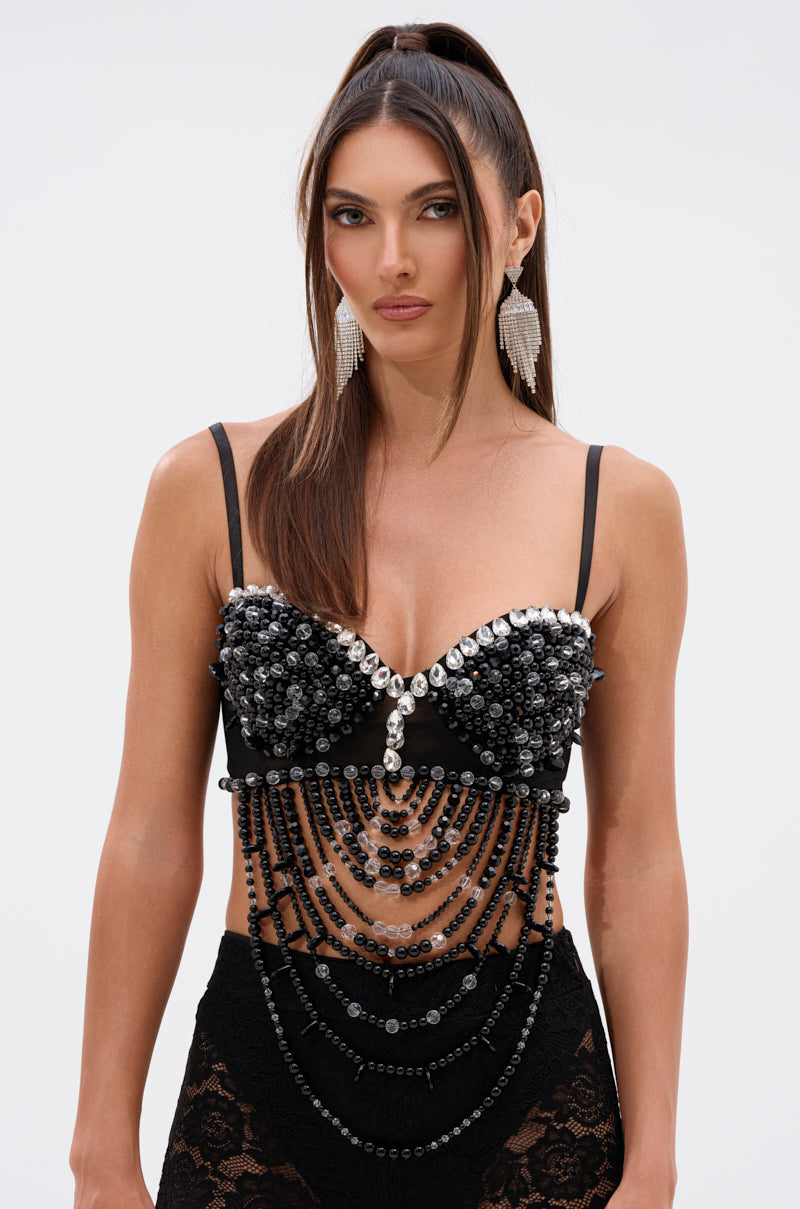 A woman with long brown hair in a high ponytail wears the EXTRA NEEDY EMBELLISHED BRALETTE, featuring draped bead details, paired with black lace bottoms and statement earrings. She stands against a plain background with a neutral expression. 1