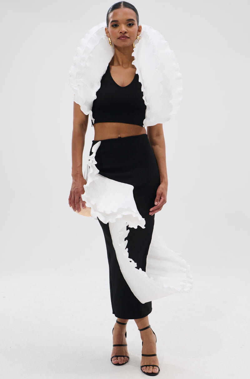 Against a plain white background, a woman wears the FASHION ICON RUFFLE BANDAGE MAXI SKIRT with bold sculptural white ruffles, paired with a matching black crop top, black heels, and hoop earrings. 0