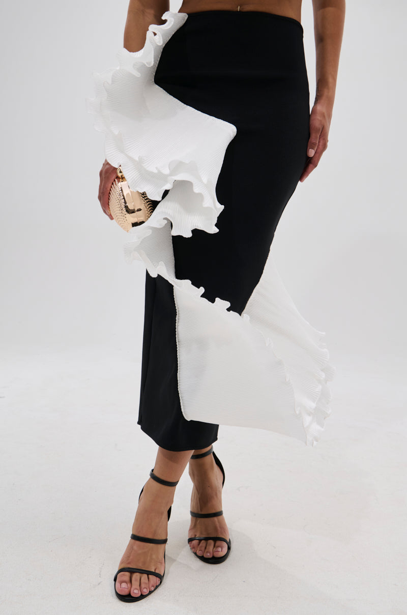 A person wearing the FASHION ICON RUFFLE BANDAGE MAXI SKIRT in black with bold white ruffle details, paired with a small metallic clutch and black strappy heels, stands against a plain white background. 1